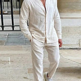 Ein Mann trägt ein naturweißes Leinen-Set, bestehend aus einem Langarmhemd mit Stehkragen und einer langen Hose, und steht in einem Innenhof mit Steinfliesen.