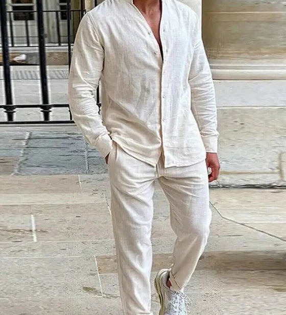 Ein Mann trägt ein naturweißes Leinen-Set, bestehend aus einem Langarmhemd mit Stehkragen und einer langen Hose, und steht in einem Innenhof mit Steinfliesen.