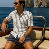 Ein Mann in einem blau-weiß gestreiften Sommer-Set, bestehend aus Kurzarmhemd und Shorts, sitzt auf einem Holzboot vor den Faraglioni-Felsen auf Capri.