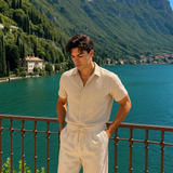Ein Mann in einem beigefarbenen, gerippten Strick-Set aus Kurzarmhemd und Shorts mit Kordelzug, steht auf einem Balkon mit Blick auf einen malerischen See und grüne Berge.
